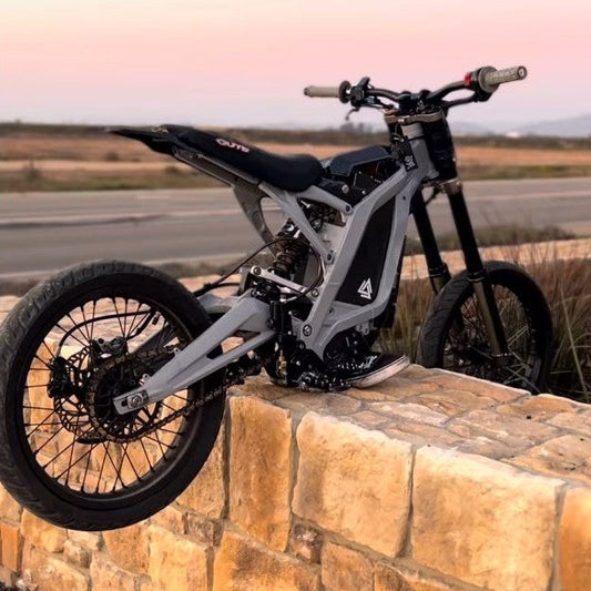 Custom Surron with a grey frame, black seat and spoked wheels, rear three-quarter view on a stone wall at sunset