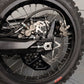 Surron Ultra Bee dual caliper bracket close-up, hexagon cutouts, foot brake caliper mounted beside the stock rear caliper