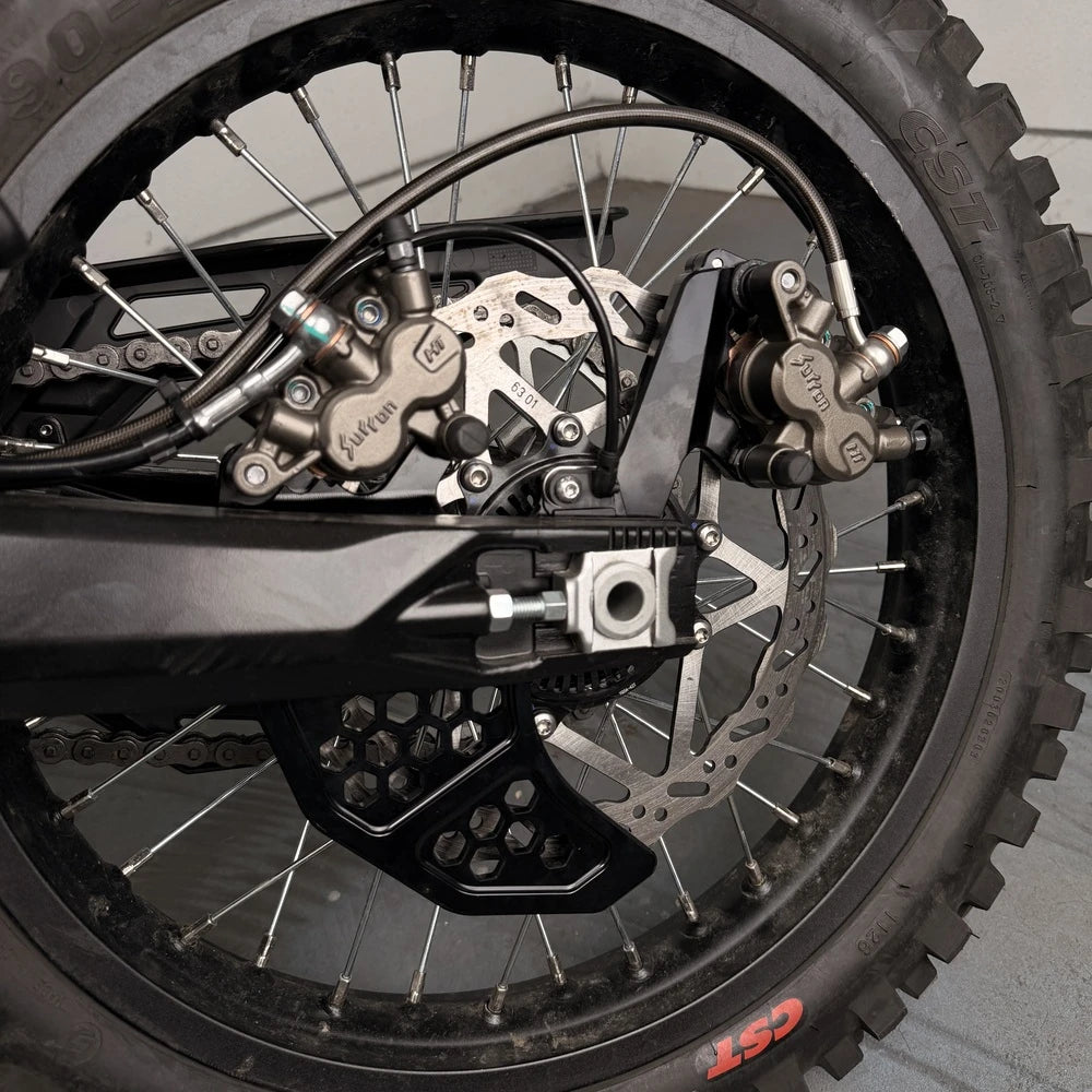 Surron Ultra Bee dual caliper bracket close-up, hexagon cutouts, foot brake caliper mounted beside the stock rear caliper